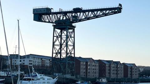 A lattice type crane beside a dock, with modern buildings behind it and large ships in the river