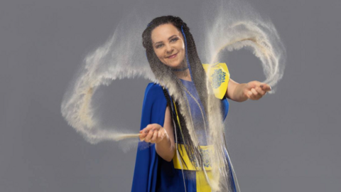 Oksana Merhut in a blue and yellow dress waving sand in front of the camera