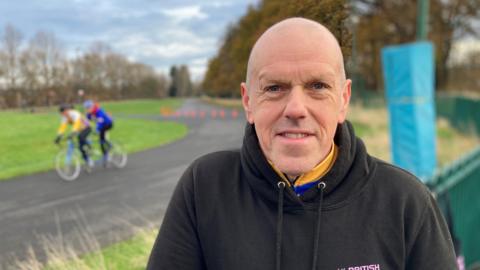 Stephen Newton is smiling at the camera while standing an a cycle track. He is bald and is wearing a black hoodie. 