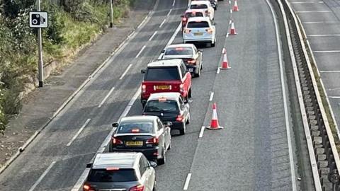 Several different coloured cars are stopped bumper-to-bumper on one side of a dual carriageway. Red and white traffic cones indicate one lane is closed. To the side you can see trees and greenery. 