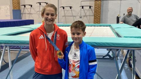 Bryony Page (left) standing in her Team GB tracksuit, with a red jacket and blue tracksuit bottoms. She is wearing her gold 2024 Olympic medal being held by Isaac, a boy wearing a blue sports jacket and a white t-shirt