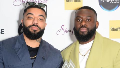 James Duncan and Fuhad Dawodu at the MOBO Awards in Sheffield in February 2024. Both men have short afro hair and beards and look seriously at the camera. James weards a dark blue shirt over a black top and has sunglasses perched on his head. Fuhad wears a pale green suit over a white top. 