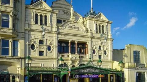 An ornate, Victorian-era theatre with a cream frontage. "Gaiety Theatre and opera house" is written in letters on the third floor, along with a greek statue of a woman holding a touch aloft. 
On the ground level there is a steel and glass overhanging shelter above the entrance. 