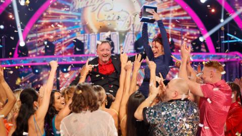 A beaming Chris McCausland, in a black blazer over a red shirt, holds his arms in the air as his dance partner, in a blue dress, holds the Glitterball Trophy aloft. The pair are surrounded by the Strictly cast who are cheering and applauding