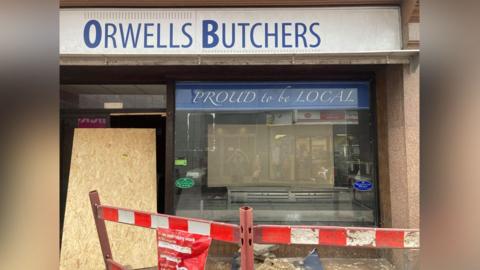 The Orwells Butchers store with red and white barriers outside and a large piece of chipboard resting against the front door. The display window is empty, but still has the "PROUD to be LOCAL" sign at the top of it.