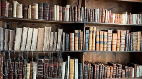 A wooden bookshelf full of old books. The majority on the left have been attached to the shelf with large metal chains, which are dangling off the edge. 