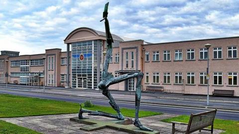 The exterior of the Isle of Man Airport, a peach coloured building with a sign that reads Ronaldsway above a glass fronted entrance, with a three legs of man statue in the foreground outside.