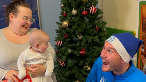 Baby Luna-Violet is in a white baby grow and is being held by her mum. She is looking at Jed Steer who wears a blue Peterborough FC branded Santa hat and is entertaining Luna-Violet by making faces. His mouth and eyes are open wide and they are both smiling. In the background there is a Christmas tree on the hospital ward. 