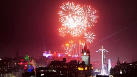 Fireworks can be seen in the sky behind the Balmoral Hotel in Edinburgh. Edinburgh Castle is lit up by the colourful fireworks and the Starflier fairground ride and Ferris Wheel are seen on the right.