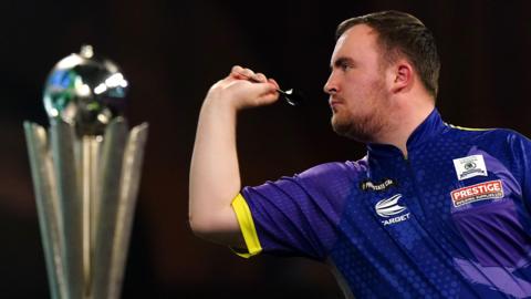 Professional darts player Luke Littler throws a dart with the championship trophy blurred in the foreground.