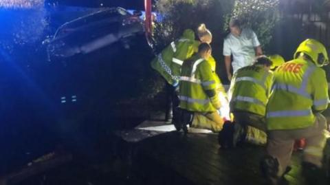 A crashed silver VW Passat sticks through a hedge. In the foreground are some firefighters