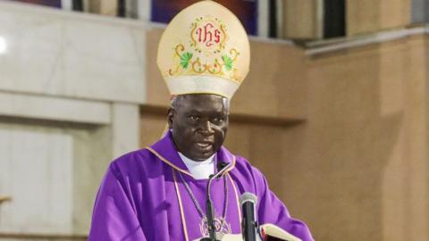 The Catholic Archbishop of Nairobi, Philip Anyolo, in purple regalia speaks before a congregation holding a microphone