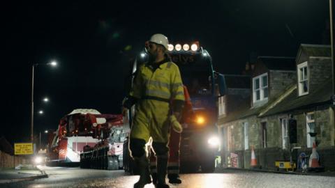 A worker walks in front of a lorry as it moves a huge power transformer past houses in a village at night