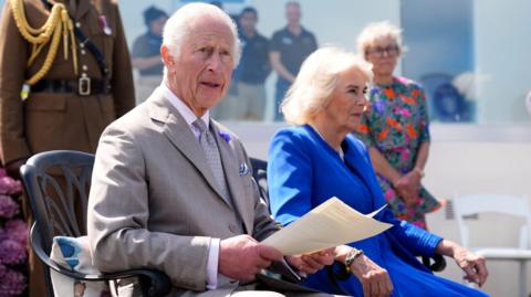 King Charles III makes a statement to members of the Alderney island with Queen Camilla, during a visit to Les Cotils at L'Hyvreuse, in Saint Peter Port, Guernsey during their two-day visit to the Channel Islands. He is sat next to the Queen. She is wearing a blue dress. The King is wearing a grey suit. 