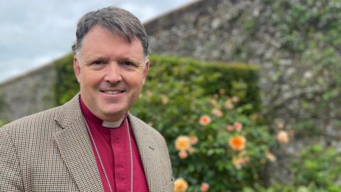 Man stands in a walled garden. hedge and roses are behind him. He wears a burgundy shirt with a cleric's dog-collar. A chain with a pendant rests on his shirt and he is wearing a brown and block tweed jacket.