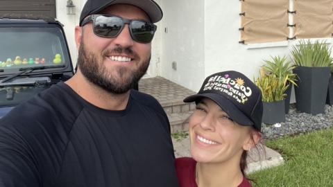 Sterling and Chynna Perkins, both wearing black caps, stand in front of a house