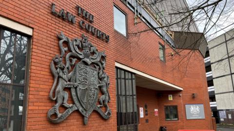 The exterior entrance of Swindon Crown Court. It is a large red brick building with a metal High Court of Justice crest attached to the wall. It reads 'The Law Courts' above it. 