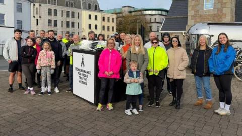 A group of about 30 people outside beside a sign saying "Remembrance for road traffic victims"