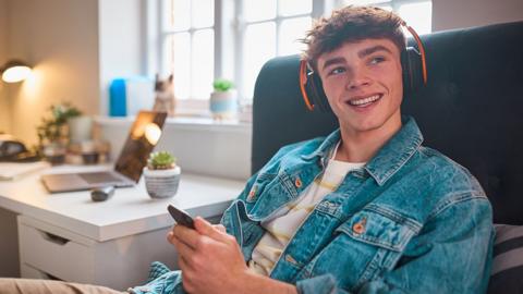 A smiling teenage student listens to a Bitesize revision podcast with headphones and a phone in their bedroom.