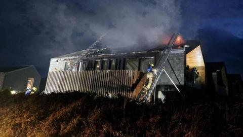 Firefighters including one climbing up a ladder trying to extinguish flames coming from the roof of a property in the early hours of the morning. Smoke is coming from the building.