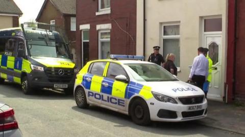 Two police cars are parked on a street outside houses. An officer in a high vis jacket speaks to a civilian woman. 