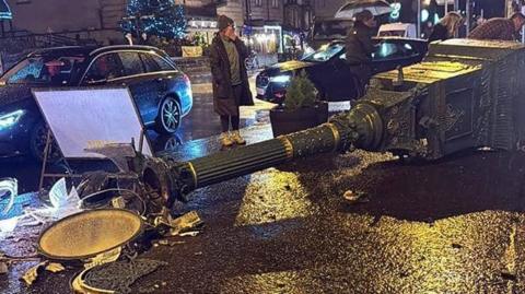 A close up of an iron clock tower which has been knocked over onto the pavement. It is about 15 feet long and is lying on its side, the clock at its head smashed. Onlookers are standing by. It is dark and wet and a christmas tree can be seen in the background.