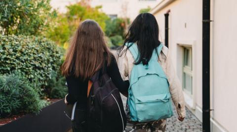 Two children walk away from the camera. The child on the left is wearing a black jumper and carrying a black rucksack, and has long brown hair. The child on the right has long dark hair and a blue rucksack, and is wearing a cream jacket. They are walking along a cobbled street with a building on their right and some bushes on their left.