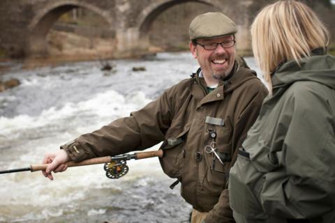 Two people fishing on the River Tweed - a man with glasses, a cap and a fishing rod talks to a blonde-haired woman