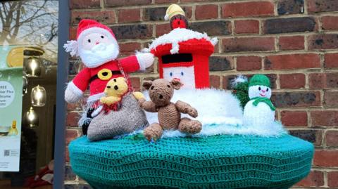 A knitted postbox topper is decorated with a Christmas scene. A snowman, teddy bear and robin on top of a knitted postbox can all be seen in a frosty scene with Santa