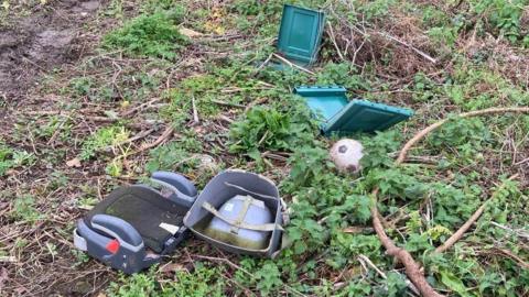 A child's car seat, a foot ball and a green plastic container have been dumped at a beauty spot. The waste items are scattered across the green foliage. Mud, sticks and stinging nettles are on the ground. 