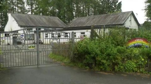 The school gates say 'Ysgol Syr John Rhys' and 'Dysg dawn daioni', with a rainbow shaped sign saying 'Ysgol Syr John Rhys'. The school is a white painted building. 