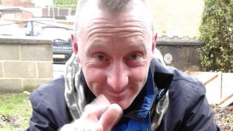 A man with short blonde hair in the middle of his head, smiles and reaches up with his right hand. He is wearing a blue coat and a wall is visible behind him.