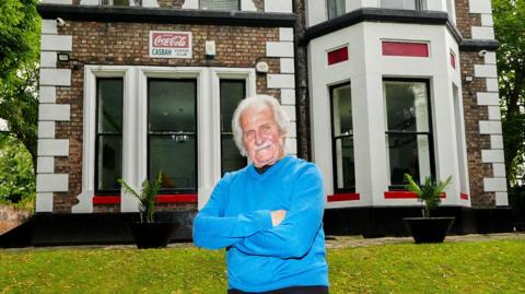 Original Beatles drummer Pete Best, 82, stood outside The Casbah Coffee Club building wearing a blue jumper with folded arms