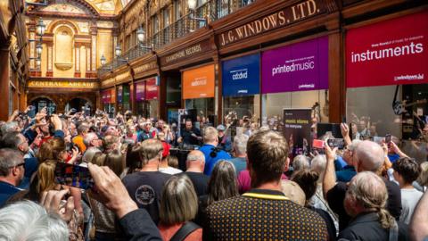 A packed Central Arcade with Lindisfarne playing outside JG Windows. People are taking pictures of the band with their phones.