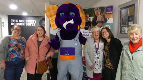 Female volunteers surround a person dressed up as a purple elephant, the charity mascot. The elephant has very large gingham checked ears. The ladies are all smiling as they pose in the Sir Bobby Robson suite.  