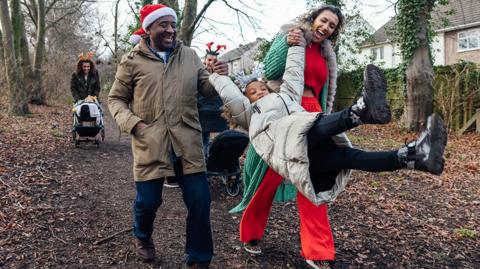 A family walking in woods near a house, all wrapped warm for winter, mum and dad swing child in the air