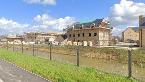 General view of the North Ely development in progress. It shows buildings with scaffolding around the outside, some have been completed and others are still under construction. The development is on the other side of a river and he sky is blue with some clouds. 