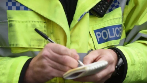 Bright yellow police jacket with the word POLICE on it. The police officer is holding a black pen and note pad