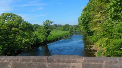 The River Ribble at Edisford Bridge, Clitheroe, showing blue skies and trees lining the river bank.