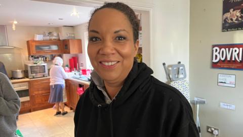 Caz Goddard is smiling to the camera, wearing a black hooded jumper. An elderly woman is pictured doing the washing up behind her.