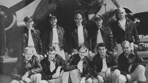 Black and white photograph of the 303rd Bomb Group in front of a B17 Bomber, also known as the Flying Fortress. There are ten men in the photograph in two rows of five. Those on the back row are standing, while those in front a crouched in a squat position. 