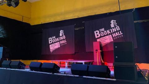 A music venue stage, two black banners at the back reading 'The Booking Hall'. The wall is yellow and there are pink lights illuminating the banners. 