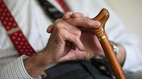 A close-up of an elderly man's hands holding a brown wooden cane. He is wearing a striped shirt with red suspenders and a dark coloured tie. 