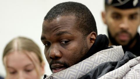 Mathias Pogba, brother of Paul Pogba, wearing a winter coat, looks to the side of the camera as he arrives to attend the verdict in his trial. Two other people are out of focus in the background behind his head. 