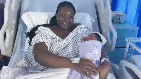 Doreen Nsiah after giving birth to baby Kylian at Walsall Manor Hospital. They are in a hospital bed and she is wearing a gown. The baby is wrapped up warm. 