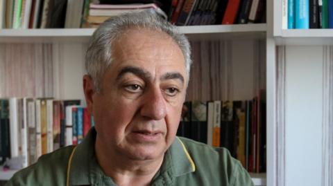 Gubad Ibadoglu sits in green t shirt in front of bookcase