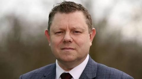 John Apter looks at the camera. He is wearing a checked suit, white shirt and tie.