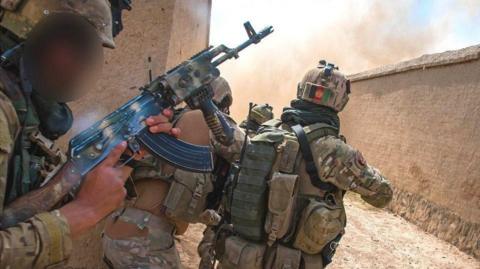 Three Afghan soldiers in camouflage gear with guns peer around the corner of a sand-coloured building, with smoke seen around the corner