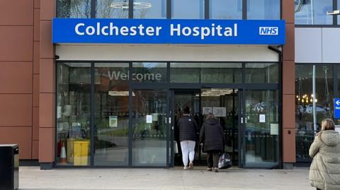 The entrance to Colchester Hospital. The name of the hospital along with the blue NHS logo sit above the doors. Patients can be seen walking through the doors.