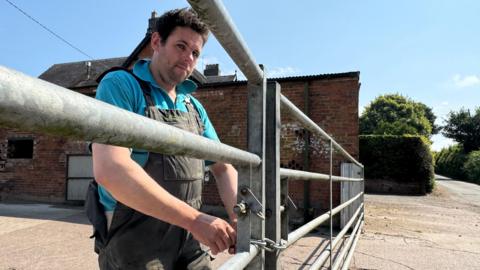 Dave Eardley is dressed in a blue t-shirt under black dungarees. He is locking a silver farm gate with a chain.